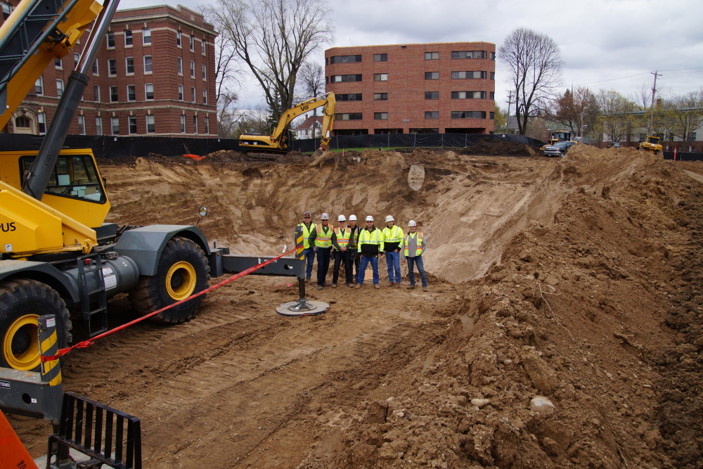 construction site with a group of people in safety vests and hardhats in the excavated hole with heavy equipment on the left