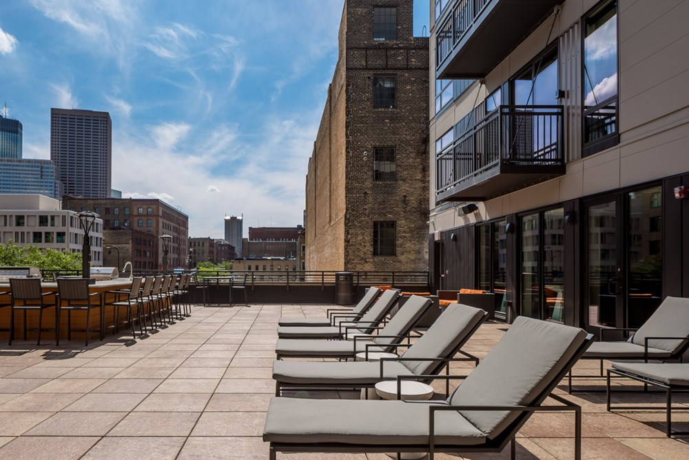 side view of a rooftop outdoor living area of an apartment building with a row of lounge chairs in front of the building on the right with a bar area with chairs on the right and a city view in the background