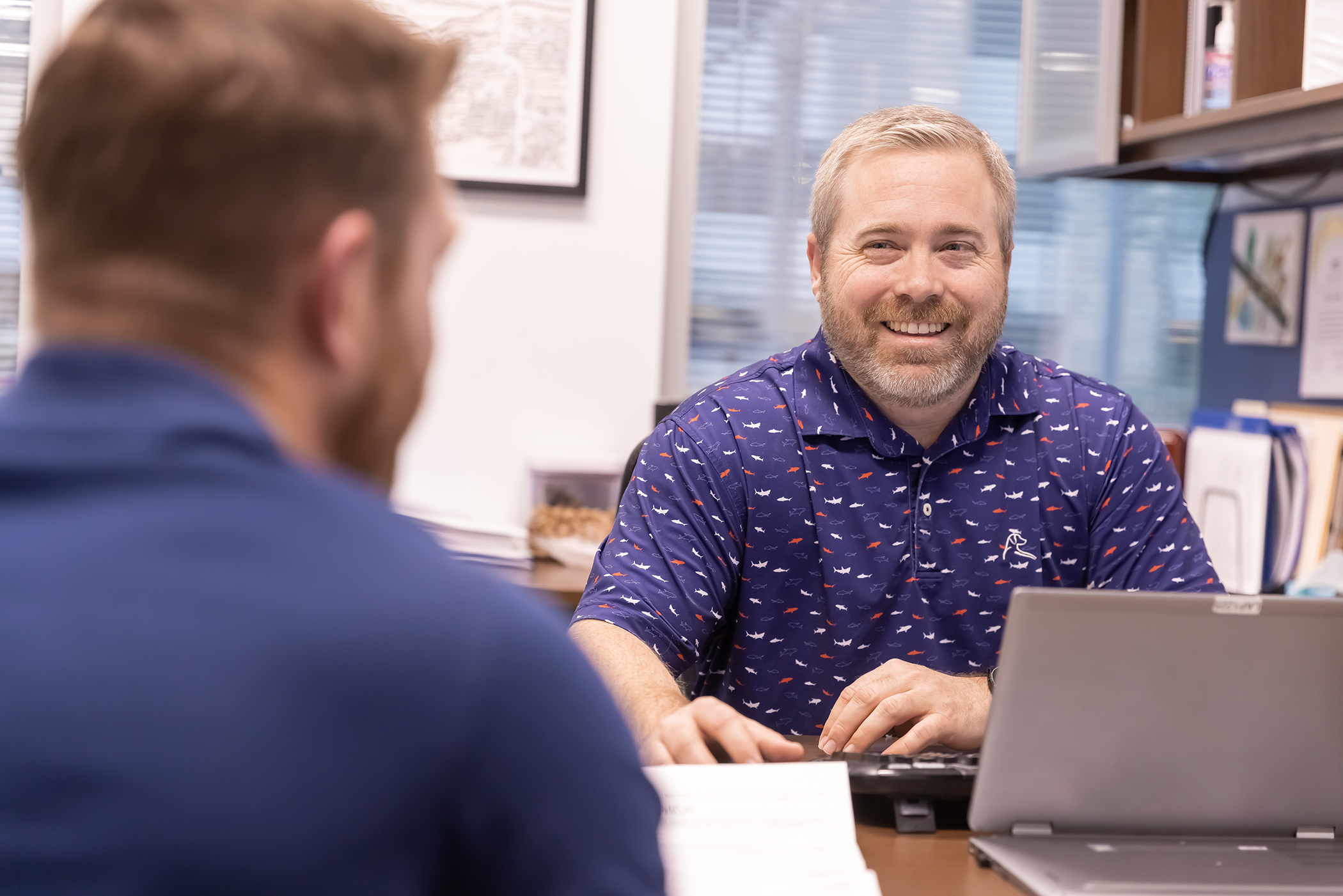 a smiling man sits at a conference room table with a laptop in front of him across from another man.